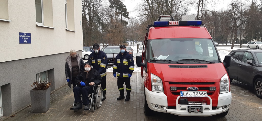 Strażacy pomagają osobie na wózku inwalidzkim dostać się do miejsca punktu szczepień przeciwko Covid-19 w tle samochód strażacki.