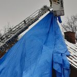 Strażak stojący w koszu drabiny mechanicznej zabezpiecza plandeką dach domu mieszkalnego.