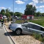 Rozbity samochód osobowy w kolorze srebrnym w rowie przy jezdni. W tle strażacy, policjanci oraz dwa samochody ratowniczo-gaśnicze i dwa ambulansy pogotowia ratunkowego.