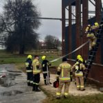 Strażacy podczas ćwiczeń przy wspinalni strażackiej. Jeden ze strażaków wchodzi po drabinie.