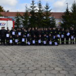 Zbiórka strażaków ochotników z dyplomami po ukończonym szkoleniu. W tle samochody strażackie.