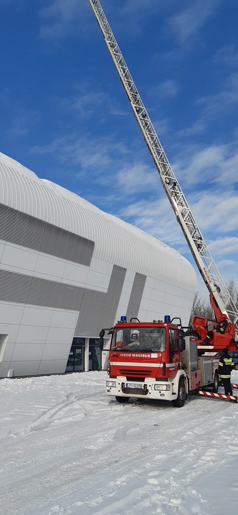 Samochód specjalny – drabina pożarnicza na tle hali sportowej.