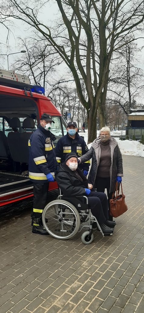 Strażacy pomagają osobie na wózku inwalidzkim dostać się do miejsca punktu szczepień przeciwko Covid-19 w tle samochód strażacki.