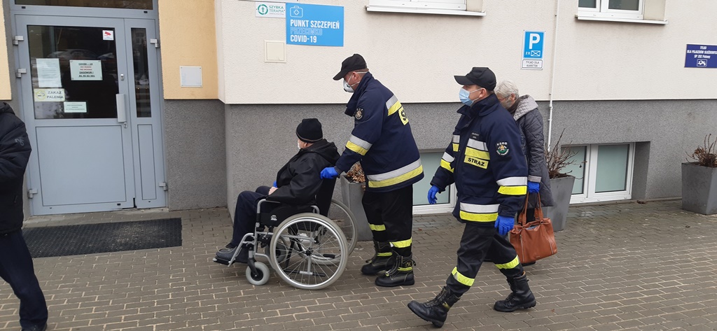 Strażacy pomagają osobie na wózku inwalidzkim dostać się do miejsca punktu szczepień przeciwko Covid-19.