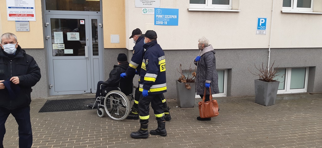 Strażacy pomagają osobie na wózku inwalidzkim dostać się do miejsca punktu szczepień przeciwko Covid-19.