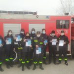 Strażacy Ochotniczej Straży Pożarnej z ulotkami w rękach na tle samochodu strażackiego.