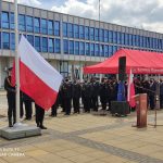 Uroczyste wciąganie flagi Polski na maszt podczas uroczystości w tle strażacy oraz zaproszeni goście.